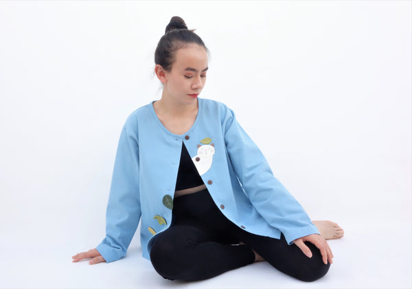 Woman sitting and wearing a blue cotton cat jacket with appliqué cat and leaves falling in the front. The jacket has buttons in the front.