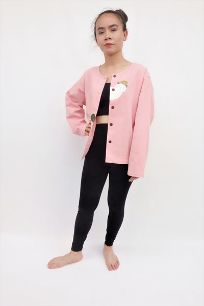 Woman standing and wearing a pink cotton cat jacket with appliqué cat and leaves falling in the front. The jacket has buttons in the front.