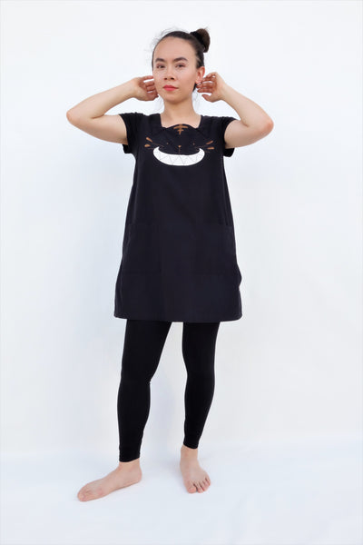 Woman wearing a cat dress, AKA pinafore dress, in black with cat appliqué, embroidery cat features, button enclosures down the left side in front full-body view.
