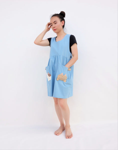 Woman wearing blue sleeveless cat dress with cats on pockets, standing side view