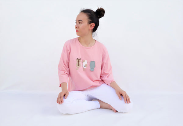 A woman is sitting casually and wearing a pink cotton cat-themed shirt with 3 appliqué cats in the front, backs facing. The shirt has three quarter sleeves and a rounded neck.