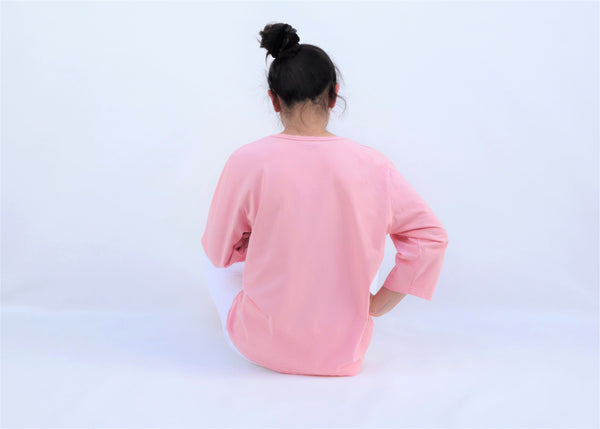A woman is sitting casually and wearing a pink cotton cat-themed shirt with 3 appliqué cats in the front, backs facing. The shirt has three quarter sleeves and a rounded neck.