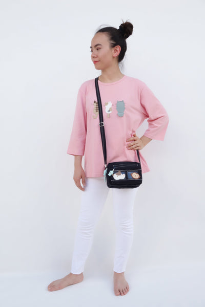 A woman is standing and wearing a pink cotton cat-themed shirt with 3 appliqué cats on the front, their backs facing. The shirt has three quarter sleeves and a rounded neck. The woman is also wearing a black cat-themed crossbody.