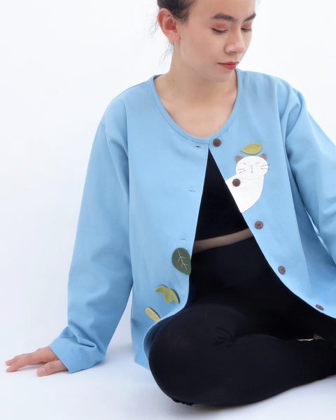 Woman sitting and wearing a blue cotton cat jacket with appliqué cat and leaves falling in the front. The jacket has buttons in the front.