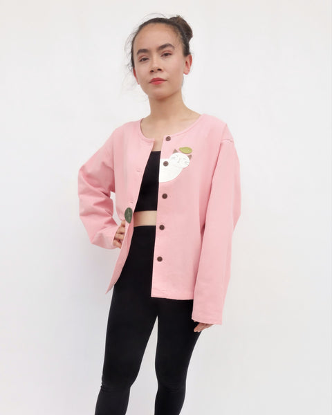 Woman standing and wearing a pink cotton cat jacket with appliqué cat and leaves falling in the front. The jacket has buttons in the front.