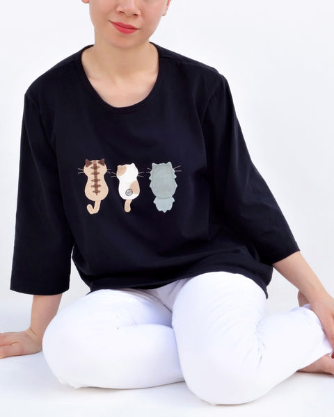 A woman is sitting casually and wearing a black cotton cat-themed shirt with 3 appliqué cats in the front, backs facing. The shirt has three quarter sleeves and a rounded neck.