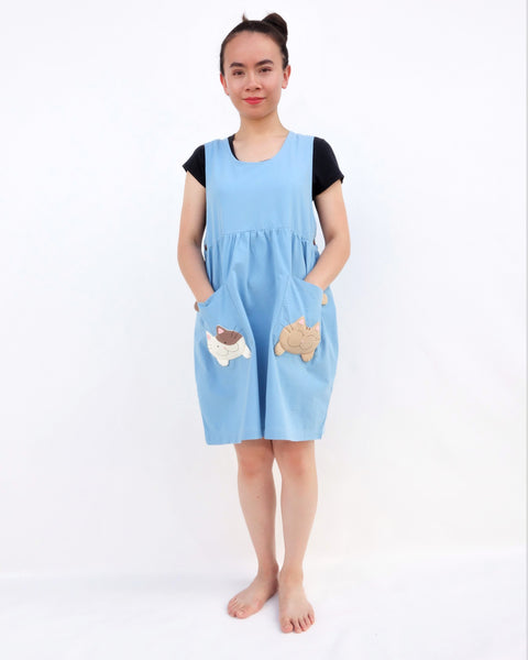 Woman wearing blue sleeveless cat dress with cats on pockets, standing and smiling