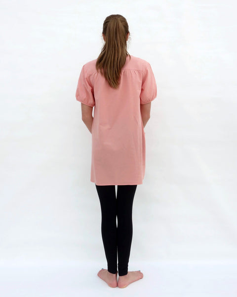 A woman standing, wearing a pink cotton cat-themed tunic dress for women with black leggings underneath, back facing. The tunic dress has puffed sleeves and slits on the side.