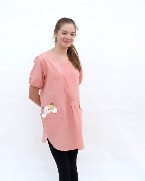 A woman standing, wearing a pink cotton cat-themed tunic dress for women with two cats/kittens on the pockets and black leggings underneath. The tunic dress has puffed sleeves and slits on the side.