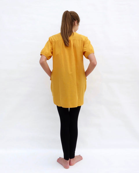 A woman standing, wearing a yellow cotton cat-themed tunic dress for women with black leggings underneath, back facing. The tunic dress has puffed sleeves and slits on the side.