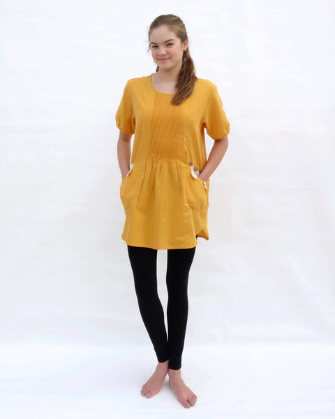 A woman with hands in the pockets, wearing a yellow cotton cat-themed tunic dress for women with two cats/kittens on the pockets and black leggings underneath. The tunic dress has puffed sleeves and slits on the side.
