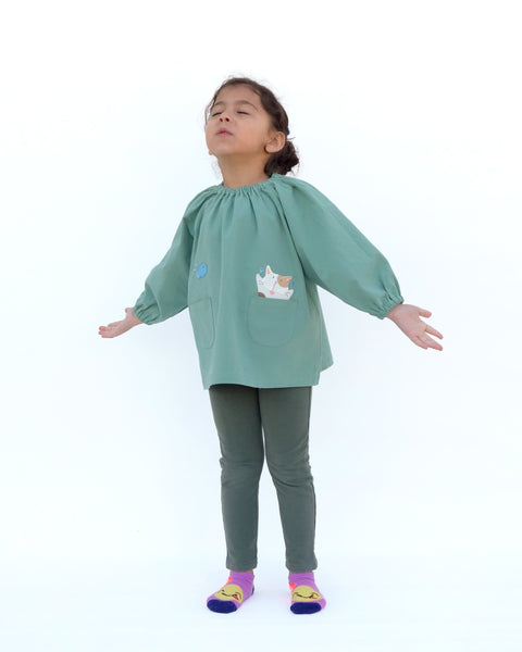 A girl wearing a loose fitting cat blouse in sage green that is light and airy with elastic around the neck, sewn-on cat appliqué, embroidery details, puffed sleeves, elastic stretch near the wrists, and two front pockets in front view