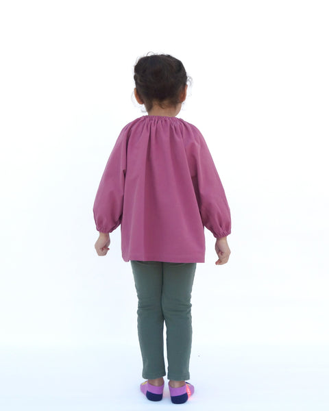 Girl wearing a loose fitting cat blouse in dull rose that is light and airy with elastic around the neck, puffed sleeves, elastic stretch near the wrists in back view.