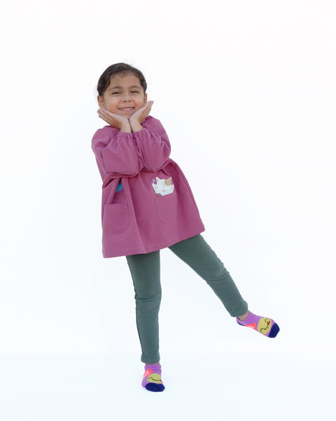 Girl wearing a loose fitting cat blouse in dull rose that is light and airy with elastic around the neck, sewn-on cat appliqué, embroidery details, puffed sleeves, elastic stretch near the wrists, and two front pockets in front view.