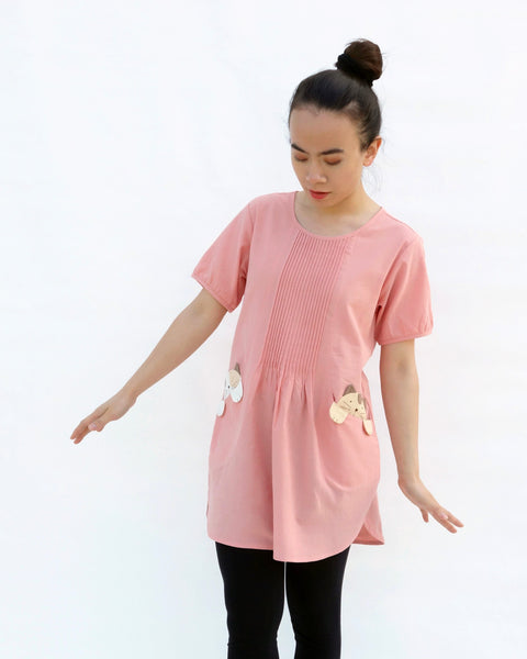 A woman standing, wearing a pink cotton cat-themed tunic dress for women with two cats/kittens on the pockets and black leggings underneath. The tunic dress has puffed sleeves and slits on the side.