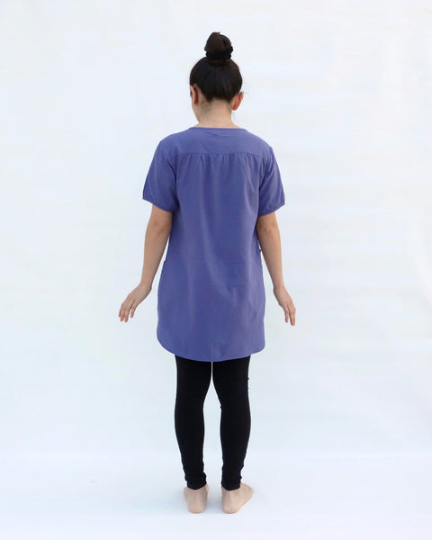 A woman standing, wearing a purple cotton cat-themed tunic dress for women with black leggings underneath, back facing. The tunic dress has puffed sleeves and slits on the side.
