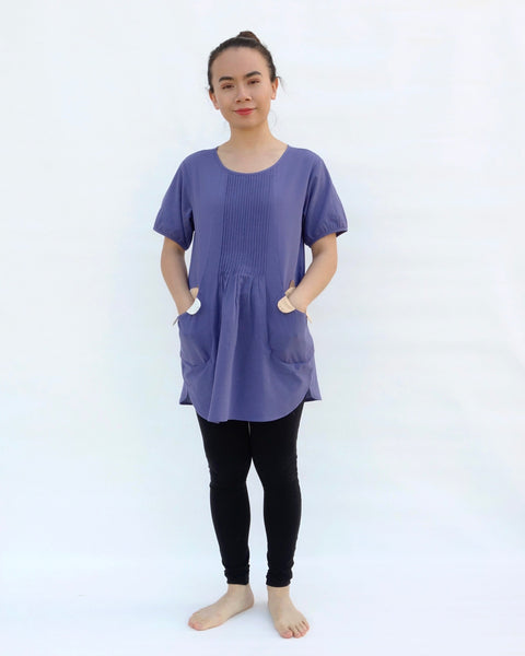 A woman with hands in pockets, wearing a purple cotton cat-themed tunic dress for women with two cats/kittens on the pockets and black leggings underneath. The tunic dress has puffed sleeves and slits on the side.