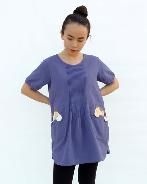 A woman with hands on hips, wearing a purple cotton cat-themed tunic dress for women with two cats/kittens on the pockets and black leggings underneath. The tunic dress has puffed sleeves and slits on the side.