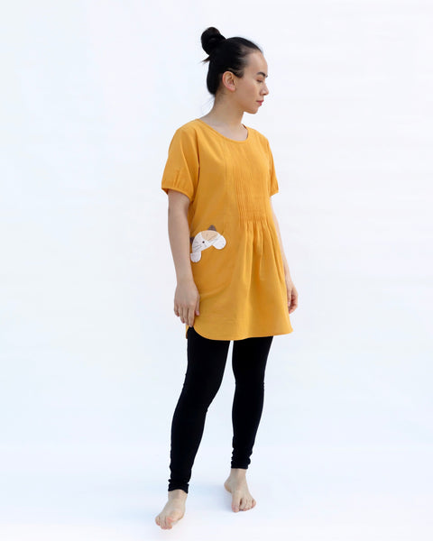 A woman with hands at her sides, wearing a yellow cotton cat-themed tunic dress for women with two cats/kittens on the pockets and black leggings underneath, side view. The tunic dress has puffed sleeves and slits on the side.