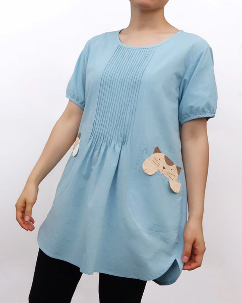 Woman wearing a sky blue cotton cat-themed tunic dress for women with two cats/kittens on the pockets and black tights leggings underneath. The tunic dress has puffed sleeves and slits on the side.
