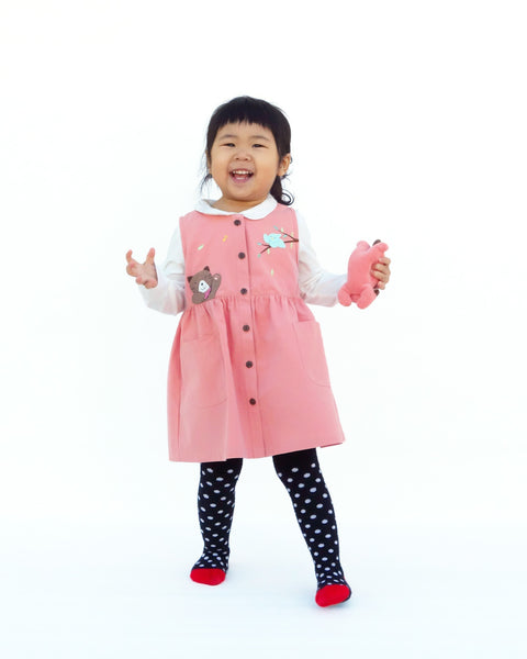 Girl wearing a cat dress in pink with high waistline, full button front, cat  appliqué, embroidery details, sleeveless, round neck opening, full-button front, two front pockets in full-body front view.