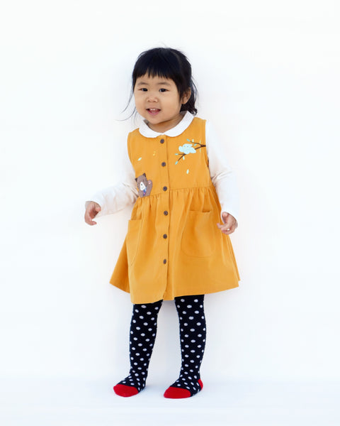 Girl wearing a cat dress in yellow with high waistline, full button front, cat  appliqué, embroidery details, sleeveless, round neck opening, full-button front, two front pockets in full-body front view.