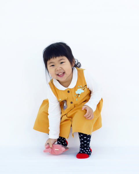 A crouching girl wearing a cat dress in yellow with high waistline, full button front, cat  appliqué, embroidery details, sleeveless, round neck opening, full-button front, two front pockets in full-body front view.