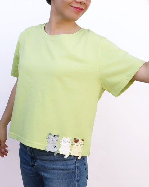 Women wearing a Cat Crop Top in green with bottom hem at the waist, three-cat appliqué on the front, one cat appliqué on the back, embroidery details, crew neck, and short sleeves in front close-up view.