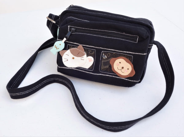 A black cat-themed crossbody purse sitting on a table. There are blue fish charms on the zippers and two rectangular PVC windows with a cat and a monkey on the front. The strap is adjustable.