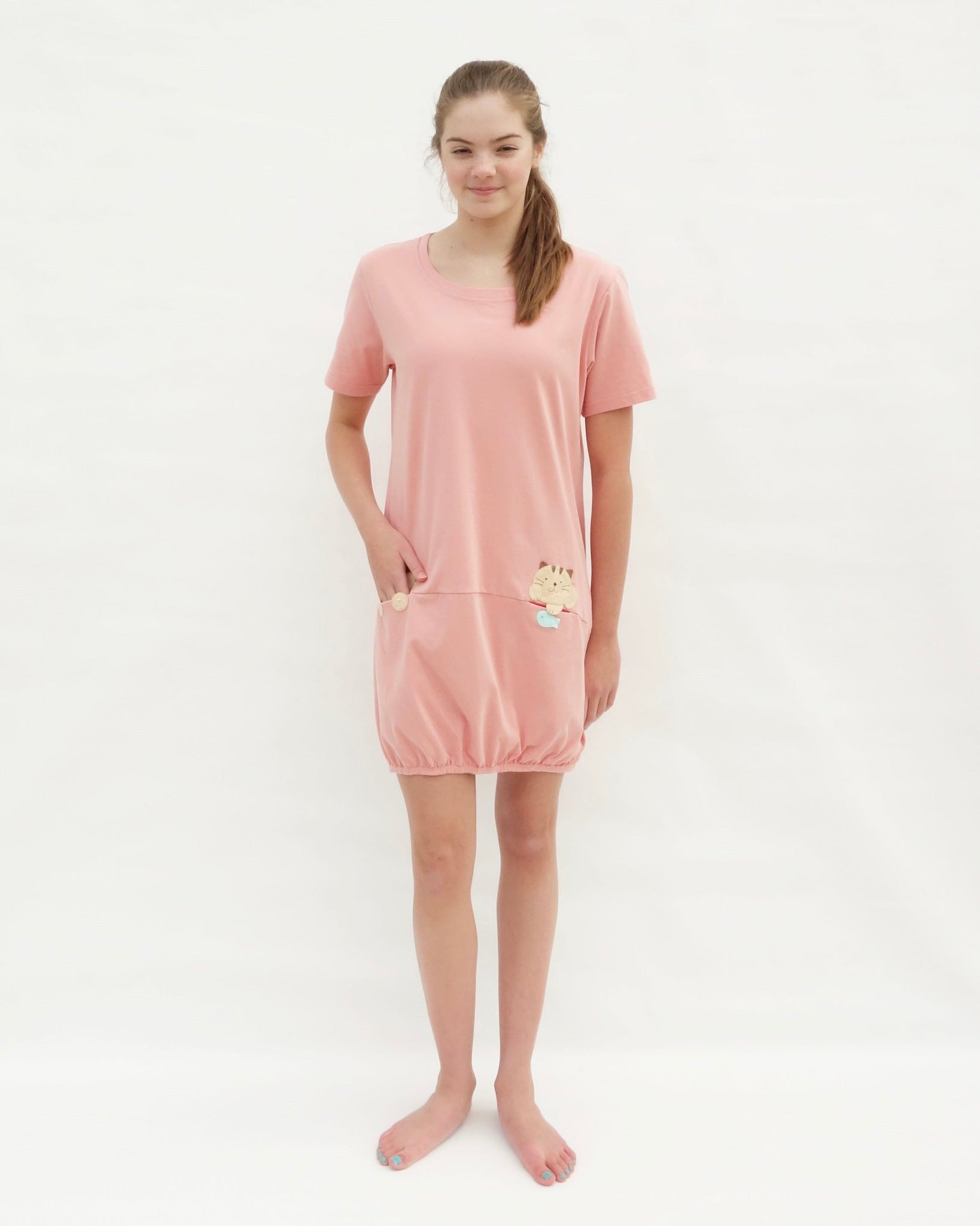 Woman wearing cat t-shirt dress in pink with cat appliqué, embroidery, front pockets, round neck opening, short sleeves, in front view.