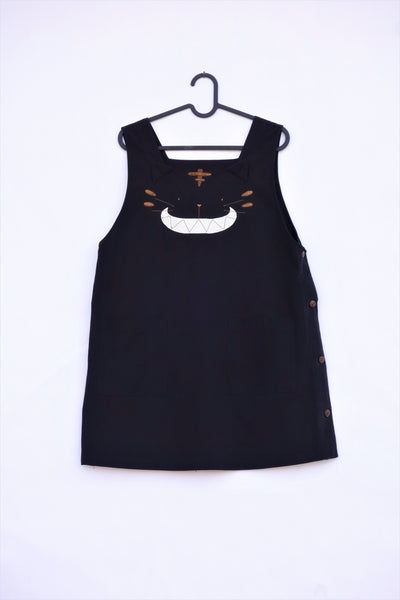 Cat Dress on a hanger, AKA pinafore dress, in black with cat appliqué, embroidery cat features, button enclosures down the left side in front view.