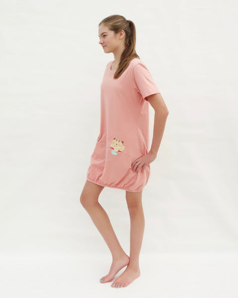 Woman wearing cat t-shirt dress in pink with cat appliqué, embroidery, front pockets, round neck opening, short sleeves, in side full-body view.