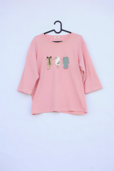 A pink cotton cat-themed shirt on a hanger with 3 appliqué cats on the front, their backs facing. The top has a rounded neck and three quarter sleeves.