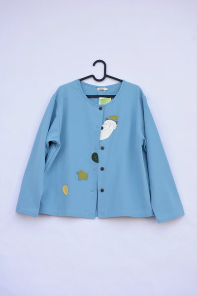 A blue women’s boxy-fit cotton cat jacket on a hanger with appliqué cat and leaves on the front. There are buttons on the jacket.