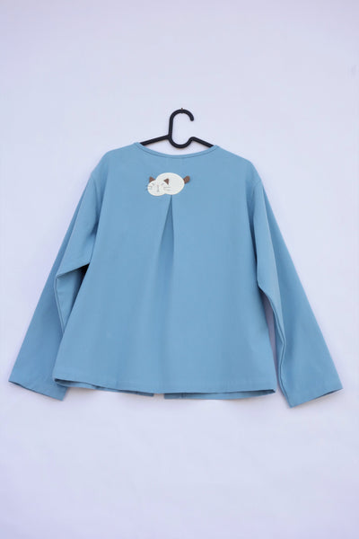 A blue womens’ boxy-fit cotton cat jacket on a hanger with an appliqué cat sleeping on the back. There is a pleat on the back of the jacket.