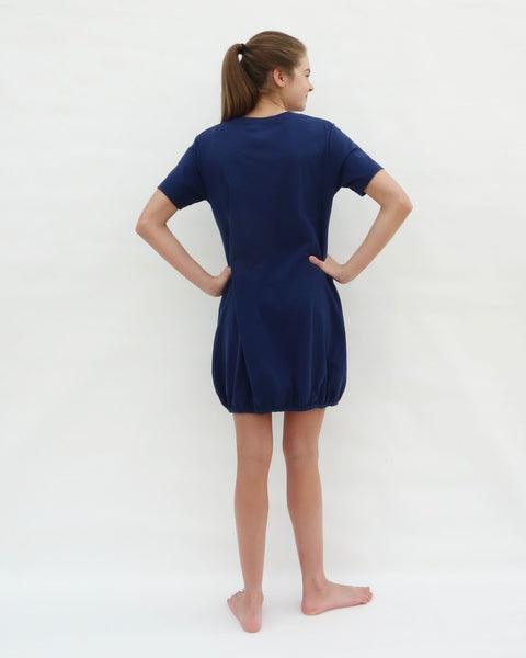 Woman wearing cat t-shirt dress in dark blue with cat appliqué, embroidery, front pockets, round neck opening, short sleeves, in back view.