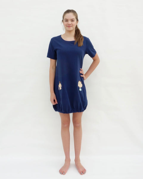 Woman with hand on hip, wearing cat t-shirt dress in dark blue with cat appliqué, embroidery, front pockets, round neck opening, short sleeves, in front full-body view.