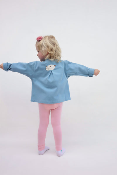 A girl is wearing a blue relaxed-fit cotton cat jacket with an appliqué sleeping cat on the back. Her arms are stretching out. There is a pleat on the back of the jacket. 