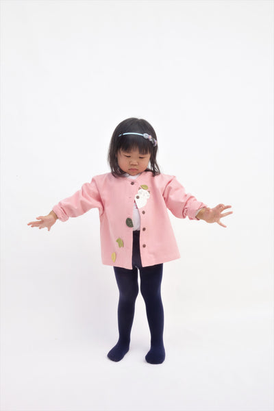 A toddler girl standing and wearing a pink relaxed-fit cotton cat jacket with an appliqué peekaboo cat and falling leaves in the front. There are buttons on the jacket and her arms are outstretched.