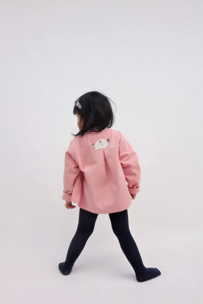 A girl is standing and wearing a pink relaxed-fit cotton cat jacket with an appliqué sleeping cat on the back. There is a pleat on the back of the jacket.