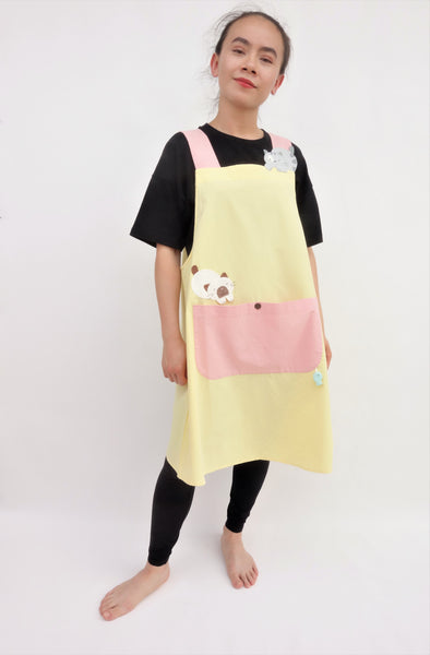 Woman wearing pink and yellow color-blocked apron with cat appliqué, large front pocket, in full-body slant view.