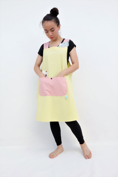 Woman wearing pink and yellow color-blocked apron with cat appliqué, large front pocket, in full-body view.