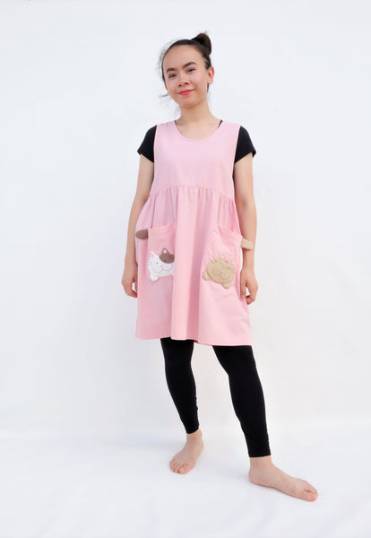 Woman wearing pink sleeveless cat dress with cats on pockets, standing and smiling
