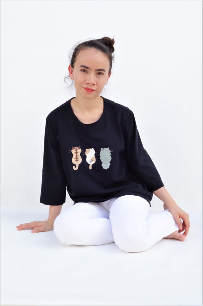 A woman is sitting casually and wearing a black cotton cat-themed shirt with 3 appliqué cats in the front, backs facing. The shirt has three quarter sleeves and a rounded neck.