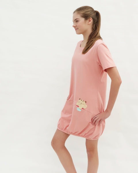 Woman wearing cat t-shirt dress in pink with cat appliqué,  embroidery, front pockets, round neck opening, short sleeves, in close-up side view.
