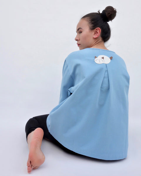 Woman sitting and wearing a blue cotton cat jacket with an appliqué cat sleeping on the back. The jacket has a pleat in the back. The woman is looking over her shoulder.
