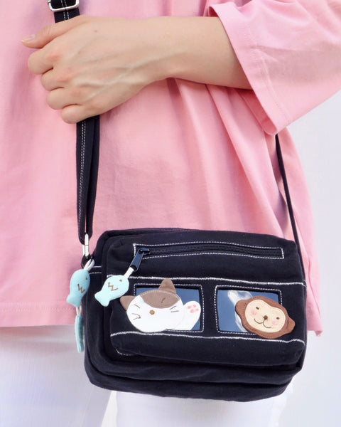 Close up of a woman holding a black cat-themed crossbody purse. There are blue fish charms on the zippers and two rectangular PVC windows with a cat and a monkey on the front. 