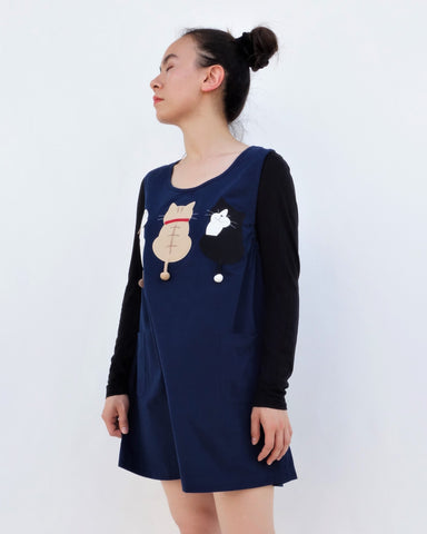 A woman standing and wearing a cat-themed, cotton, dark blue shirt/mini A-line dress/top/tunic with three appliqué cats on the front