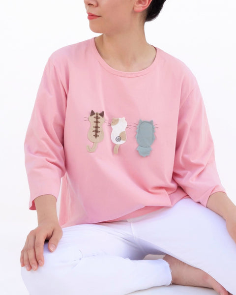 A woman is sitting casually and wearing a pink cotton cat-themed shirt with 3 appliqué cats in the front, backs facing. The shirt has three quarter sleeves and a rounded neck.