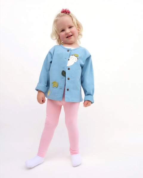 A girl standing, smiling, and wearing a blue relaxed-fit cotton cat jacket with an appliqué peekaboo cat and falling leaves in the front. There are buttons on the jacket. 